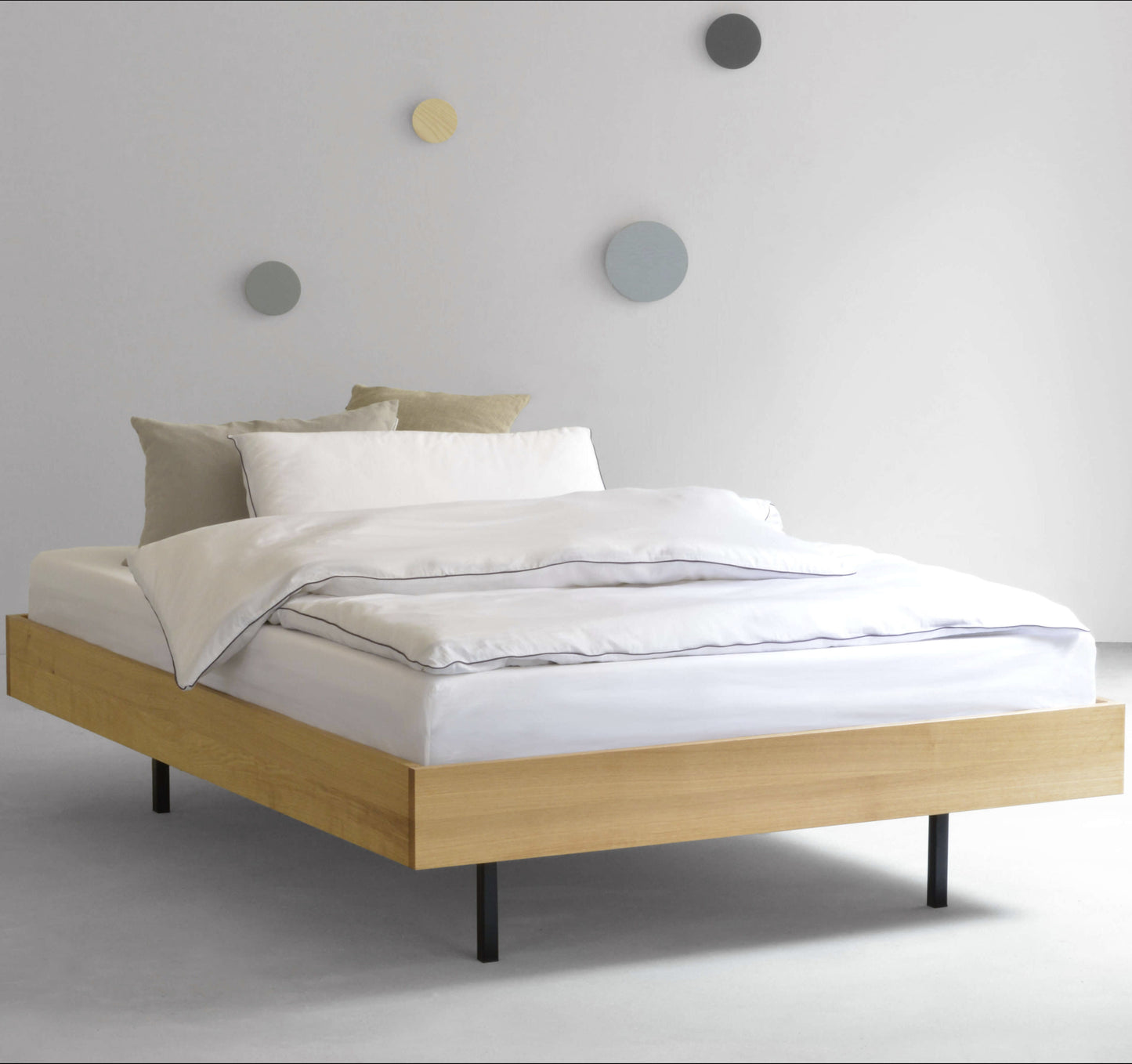 Eichenbett mit schwarzen Stahlfüßen steht auf einem hellen Boden im Hintergrund sieht man farbige, runde Haken an der wand