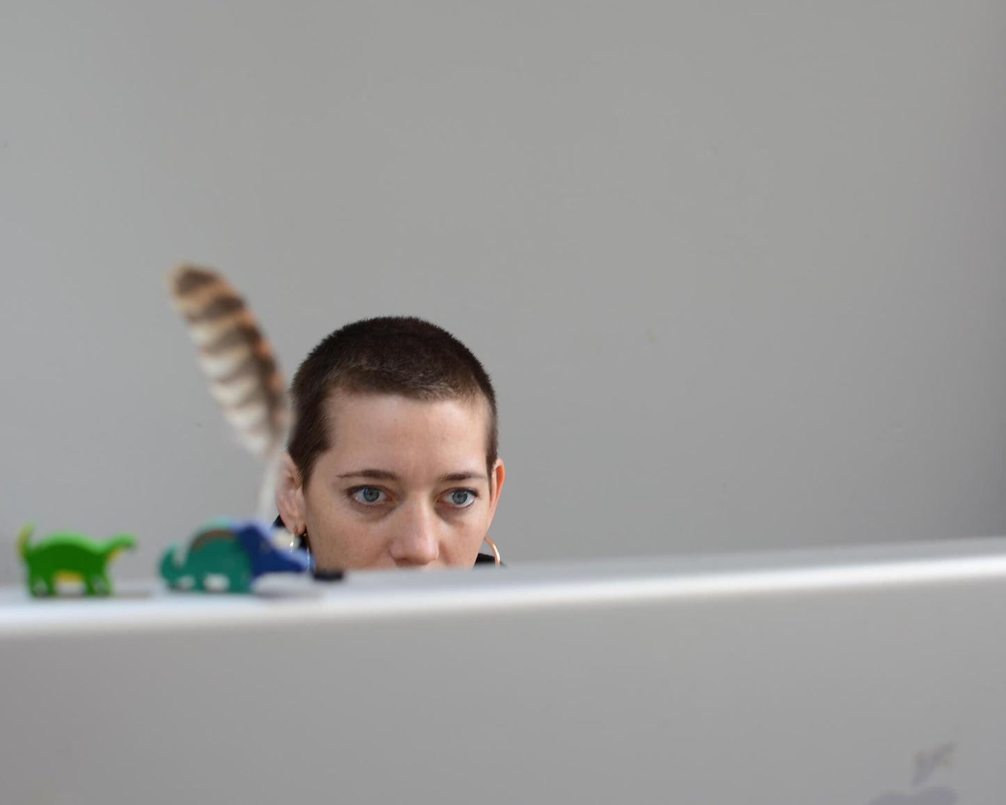 Kopf einer Person, die hinter einem Monitor nur halb zu sehen ist – dazu eine Feder und Spielzeug im Hintergrund
